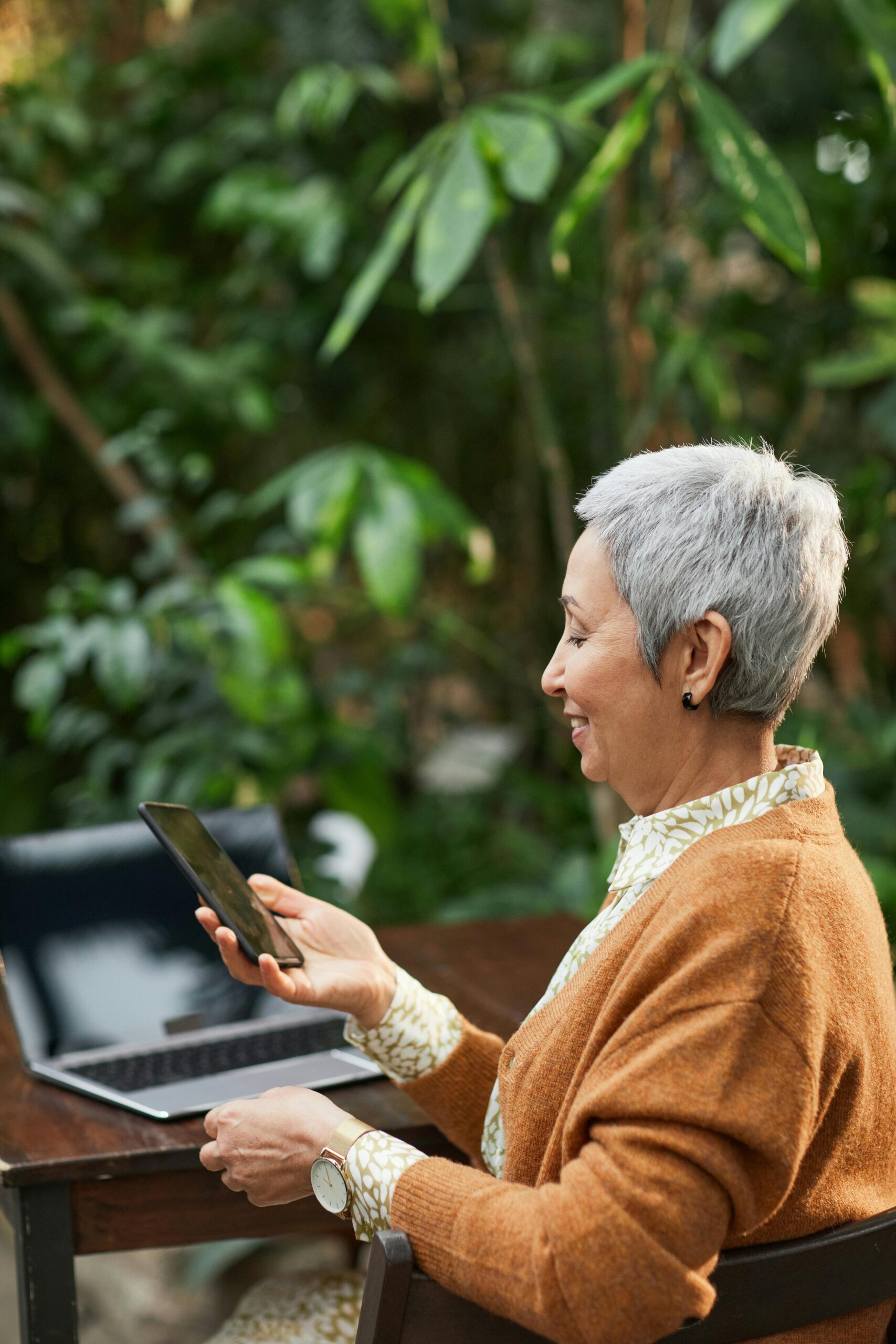 Elderly woman smiling while using a smartphone at an outdoor table, surrounded by lush greenery.